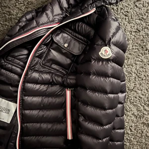 Svart dunjacka från Moncler - Snygg svart dunjacka från Moncler (köptes på plick) med huva och dragkedja i rött, vitt och blått. Jackan har två bröstfickor med knappar och ett quiltat mönster som ger en stilren look. Perfekt för kyliga dagar. Storlek 3 och fullt fungerande nfc scann. Hör av er vid fler frågor och funderingar🥂🙌