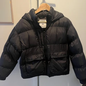 Svart dunjacka från Hoodrich - Säljer en svart dunjacka från Hoodrich i storlek XS. Jackan har en pufferdesign med huva och dragkedja framtill. Den har också praktiska fickor med knappar och ett broderat Hoodrich-märke på ryggen.