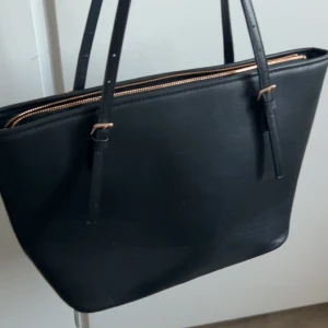 Svart axelväska med dragkedja - Snygg svart axelväska i syntetmaterial med praktisk dragkedja upptill. Perfekt för att bära med sig det nödvändigaste i stil.