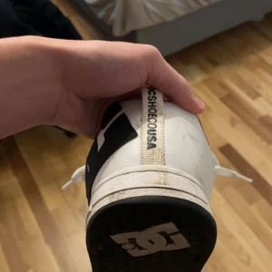 Vita och svarta sneakers från DC Shoes - Säljer ett par vita sneakers från DC Shoes med svart logga på sidan. Skorna har snörning och är i bra skick, perfekta för vardagsbruk. De har en klassisk design med en bekväm passform.