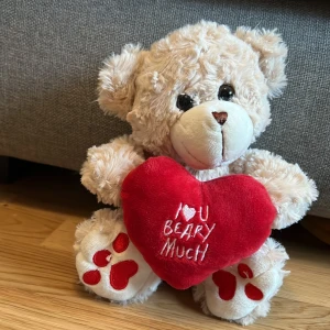 Mjuk nallebjörn med hjärta - Söt beige nallebjörn som håller ett rött hjärta med texten 'I love you beary much'. Perfekt som present eller dekoration. Nallen har vita tassar med röda detaljer och är gjord av mjukt plyschmaterial.