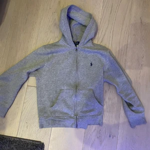 Grå hoodie från Ralph Lauren - Säljer en stilren grå hoodie från Ralph Lauren med dragkedja och klassisk logga på bröstet. Tröjan har en bekväm huva och praktiska fickor framtill. Perfekt för en avslappnad look. Den är i super bra skick nästan som att den aldrig används