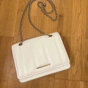 Vit axelväska från Pull&Bear - Snygg vit axelväska från Pull&Bear med kedjerem och stilren design. Väskan har en elegant gulddetalj på framsidan och är perfekt för att bära dina nödvändigheter. Den har ett praktiskt innerfack och stängs med en tryckknapp. På sista bilden ser du dock att den har en liten fläck på insidan, men det är inget man tänker jättemycket på! 💞