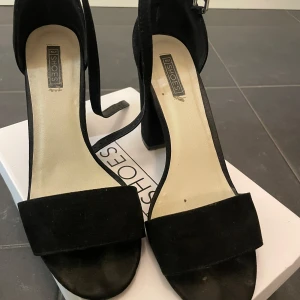 Svarta klackskor från Nelly - Snygga svarta klackskor i mocka. De har en elegant design med öppen tå och en bred rem över foten. Skorna har en hög klack och en justerbar rem runt ankeln för perfekt passform. Perfekta för festliga tillfällen!