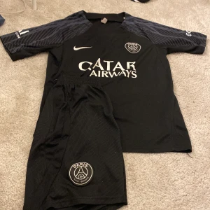 Svart PSG fotbollströja - Säljer en snygg svart PSG fotbollströja från Nike. Den har korta ärmar och ett stilrent märke på bröstet. Perfekt för alla fotbollsfans som vill visa sitt stöd för Paris Saint-Germain. Tröjan har också Qatar Airways som sponsorlogga. Passar perfekt till både träning och vardag!