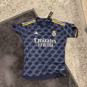 Real Madrid fotbollströja med Vini Jr. 7 - Säljer en ny Real Madrid fotbollströja från Adidas med Vini Jr. och nummer 7 på ryggen. Tröjan är mörkblå med ett subtilt mönster och har gula detaljer på ärmarna. Den är kortärmad och har klubbens emblem broderat på bröstet. Perfekt för fans som vill visa sitt stöd!