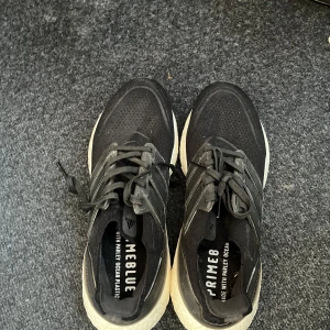 Svarta Adidas sneakers - Säljer ett par svarta Adidas sneakers med vit sula och snörning. Skorna är märkta med 'Primeblue' och är tillverkade med återvunnen plast. Perfekta för en sportig look.