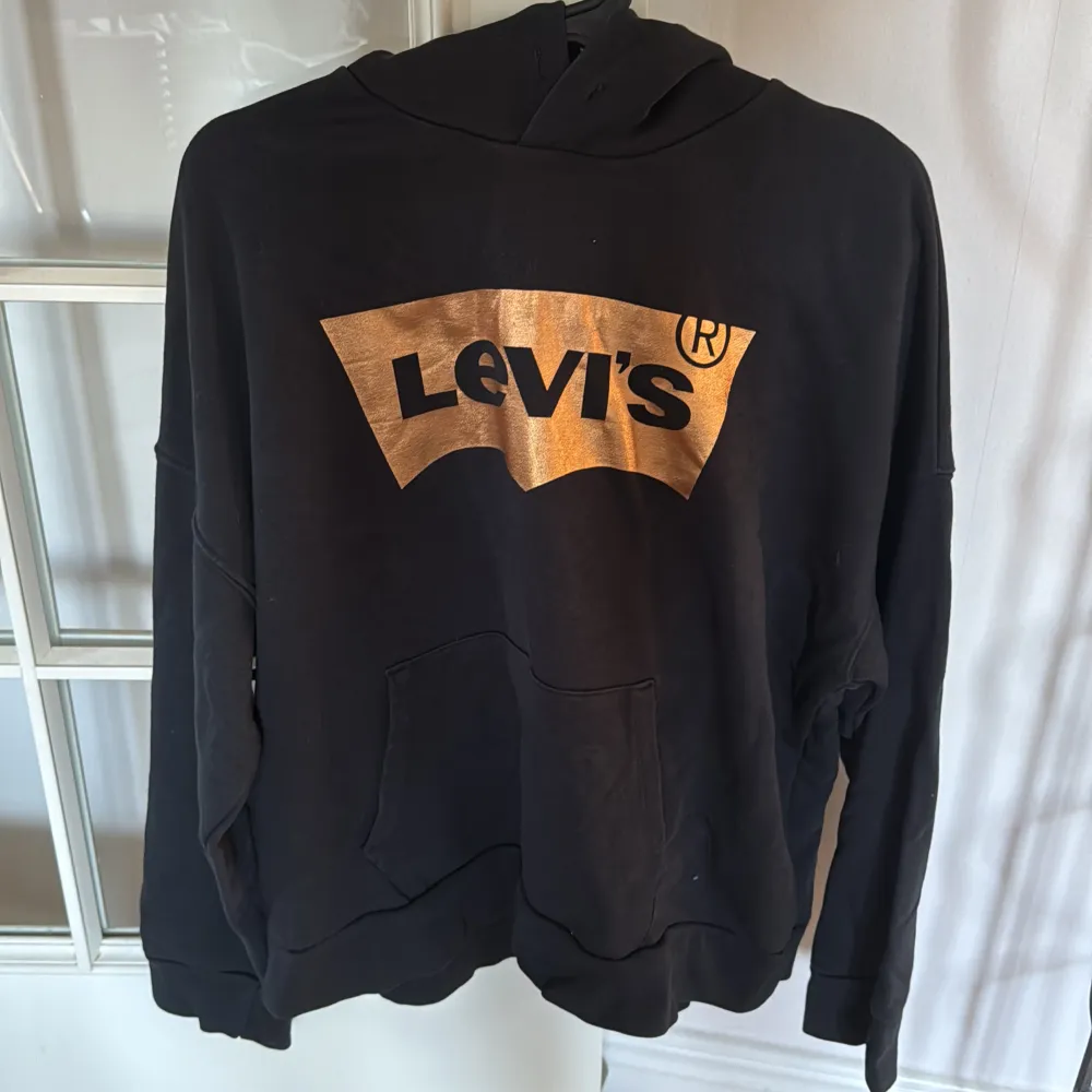 Snygg svart hoodie från Levi's med en stor, glittrig logga i guld på bröstet. Tröjan har en klassisk känguruficka och en bekväm huva. S men passar M oxå. Tröjor & Koftor.