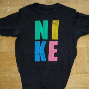 Vintage Svart t-shirt från Nike från 90-talet - Vintage svart t-shirt från Nike från 90-talet, färgglad text i grönt, gult, blått och rosa. Den har korta ärmar och en klassisk rund halsringning. Perfekt för en avslappnad stil. 