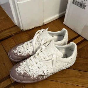 Maison Margiela sneakers i vitt och rosa - Säljer ett par unika Maison Margiela sneakers i vitt med rosa detaljer. Skorna har en cool, sliten look med färgstänk och snörning. Perfekta för dig som vill sticka ut med en konstnärlig stil. Använda men fortfarande i bra skick.