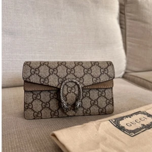 Gucci axelväska med monogram - Säljer en snygg Gucci axelväska i mycket bra skick. Väskan har det klassiska Gucci-monogrammet i beige och brunt, med en elegant kedjerem och en dekorativ spänne framtill. Perfekt för både vardag och fest! 👜✨
