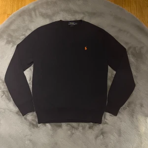 Marinblå tröja från Ralph Lauren - Säljer en stilren marinblå tröja från Ralph Lauren i bomull. Den har en klassisk v-ringning och det ikoniska broderade logotypen i orange på bröstet. Perfekt för både vardag och fest! 