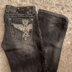 Grå bootcut jeans med slitningar från Miss Me - Säljer ett par grå jeans från Miss Me i storlek 28. De har slitningar på framsidan och snygga broderade detaljer på bakfickorna. Knappen på framsidan är borta och var även det när jag köpte dem, köpte dem scondhand men har själv aldrig använt dom. 