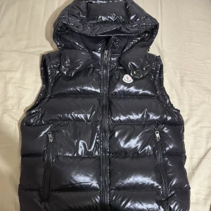 Svart Moncler väst  - Säljer en snygg svart Moncler väst i mycket bra skick, har använts bara 2 gånger. Västen har en glansig yta med dragkedja framtill och två praktiska fickor. Perfekt för höst och vinter, håller dig varm och stilren. Passar både till vardags och festliga tillfällen.