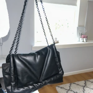Svart axelväska med kedja - Snygg svart axelväska i skinn med kedjedetaljer. Perfekt för både vardag och fest! Väskan har en stilren design och är rymlig nog för alla dina nödvändigheter. Den är i mycket bra skick och redo för nya äventyr.
