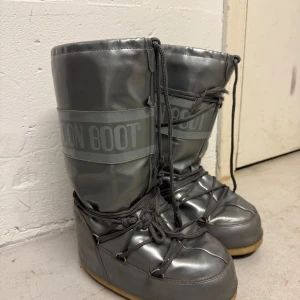 Grå Moon Boots - Säljer ett par grå Moon Boots i bra skick. Har ett litet hål i hälen. De är perfekta för vintern med sitt varma och vadderade material. Skorna har snörning och en cool, futuristisk look. Perfekta för att hålla fötterna varma och stiliga i snön! Strl 39