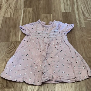 Rosa blommig klänning för barn - Söt rosa klänning från Name It med små blommor och korta ärmar. Klänningen har en smockdetalj framtill och knappar i ryggen för enkel påklädning. Perfekt för varma dagar!