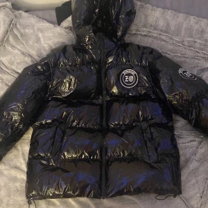 Svart pufferjacka med avtagbara ärmar - Säljer en svart, glansig pufferjacka Jackan har avtagbara ärmar och en stor logga på ryggen. Perfekt för vintern med sin varma och quiltade design. Den har en dragkedja framtill och en huva för extra skydd mot kylan. Supercool och mångsidig stil! 🖤den är helt ny men bara pröv testa. Storlek S men passar med M