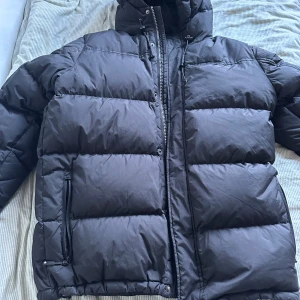 Svart pufferjacka från Cross Sportswear - Säljer en svart pufferjacka från Cross Sportswear i bra skick. Jackan har en dragkedja framtill och är perfekt för kalla vinterdagar. Den är långärmad och har en normal passform. Supermysig och varm! Dock har ena knappen lossnat och knappen till snöret där man spänner luvan, men det gör ingen skillnad på jackan det syns inte endån.