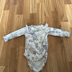 Blommig långärmad body från Newbie - Söt långärmad body från Newbie med ett blommigt mönster i lila och beige toner. Bodyn har volangdetaljer vid axlarna och är perfekt för de minsta. Tillverkad i mjukt material för extra komfort.