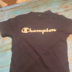 Säljer en svart t-shirt från Champion i bomull. Den har korta ärmar och en stor logga på framsidan. Lappen är avklippt men storleken är 122/128