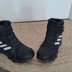 Svarta kängor från Adidas - Säljer ett par svarta kängor från Adidas i nyskick. De har en cool design med vita detaljer och snörning framtill. Perfekta för höst och vinter med en robust sula som ger bra grepp. De har också en praktisk rem med kardborre för extra stöd.