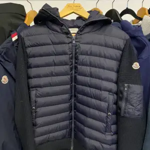 Säljer en snygg mörkblå dunjacka från Moncler. Jackan har en quiltad design med stickade ärmar och huva. Den har en dragkedja framtill och en praktisk ficka med dragkedja på ärmen. Perfekt för kyliga dagar!