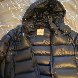 Mörkblå  pufferjacka från Moncler - Säljer en snygg och varm svart pufferjacka från Moncler. Jackan har en glansig finish och är perfekt för kalla vinterdagar. Den har en praktisk huva och stängs med dragkedja. Perfekt för att hålla stilen samtidigt som du håller dig varm!