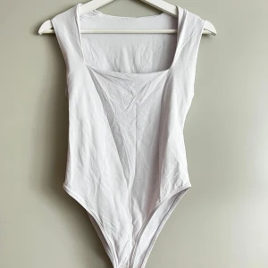 Vit ärmlös bodysuit - Säljer en stilren vit bodysuit i mjukt material. Den är ärmlös och har en enkel design som passar perfekt till både vardag och fest. Bodysuiten är i nyskick och har en tight passform.
