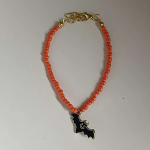 Armband med fladdermusberlock - Säljer ett coolt armband med orange pärlor och en svart fladdermusberlock. Perfekt för Halloween eller när du vill ha en unik accessoar. Armbandet har guldfärgade detaljer och ett justerbart lås.