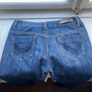 Blå jeans från Miss Two - Säljer ett par snygga low waisted bootcut jeans från Miss Two. De har en klassisk femficksdesign med coola broderade detaljer på rumpan. Jeansen har en knapp och dragkedja framtill. 🤗🤗 PRIS GÅR ATT DISKUTERA