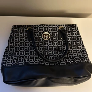 Svart och vit Tommy Hilfiger handväska - Säljer en nästan oanvänd svart och vit Tommy Hilfiger handväska. Väskan har svarta handtag och detaljer i guld. Den är rymlig och perfekt för vardagsbruk. Insidan har en dragkedja för extra säkerhet.