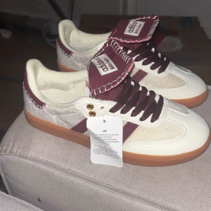 Adidas sneakers med vinröda detaljer - Snygga Adidas sneakers i vitt med vinröda detaljer och klassiska tre ränder. Skorna har en gummisula i ljusbrun färg och snörning framtill. Perfekta för en stilren look.