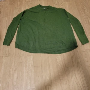 Grön stickad tröja från Holly & Whyte - Säljer en snygg grön stickad tröja från Holly & Whyte. Tröjan har en enkel design med långa ärmar och ribbade kanter. Perfekt för kyligare dagar och ger en stilren look.