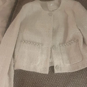 Vit kavaj med pärldetaljer - Elegant vit kavaj med glittriga pärlknappar och dekorativa pärlor på fickorna. Den har en klassisk passform och är tillverkad i ett strukturerat tyg som ger en lyxig känsla. Perfekt för att lyfta vilken outfit som helst. Storlek M anser att den även passar S, aldrig använd🩷