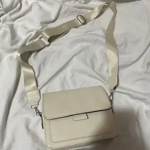 Snygg beige axelväska med en justerbar rem för perfekt passform. Väskan har en stilren design med ett lock och en praktisk storlek för vardagsbruk. Perfekt för att bära dina nödvändigheter med stil.