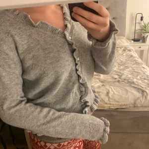 Grå kofta med volangdetaljer - Säljer en mysig grå kofta med volangdetaljer längs knäppningen. Koftan har långa ärmar och ribbade kanter vid ärmslut och nederkant. Perfekt för kyliga dagar! Vet ej vart den är ifrån.