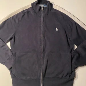 Mörkblå tröja med dragkedja från Ralph Lauren - Snygg mörkblå tröja från Ralph Lauren med dragkedja och klassisk logga på bröstet. Tröjan har långa ärmar och ribbade muddar vid ärmslut och nederkant. Perfekt för en stilren look.