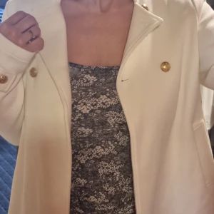Vit kappa från Molly Bracken - Elegant vit kappa från Molly Bracken med guldknappar och stilren design. Perfekt för att lyfta din outfit med en touch av klass. Kappan har en slits baktill och är långärmad.