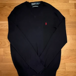 Ralph lauren tröja - Säljer en stilren marinblå tröja från Polo Ralph Lauren med röd broderad logga på bröstet. Tröjan har en v-ringad design och är tillverkad i bomull, vilket gör den både bekväm och snygg. Perfekt som ett varmt o snyggt lager under jackan. Mvh C.W Closet