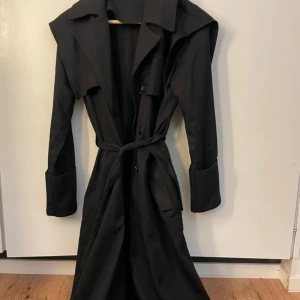 Svart trenchcoat med bälte - Elegant svart trenchcoat med knappar och bälte i midjan. Jackan har långa ärmar och en klassisk krage, perfekt för en stilren look.