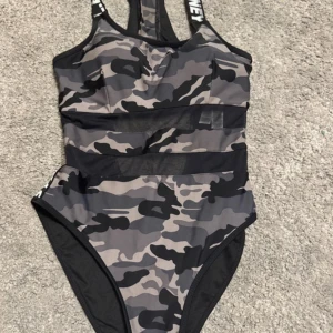 Kamouflagemönstrad baddräkt - Snygg kamouflagemönstrad baddräkt med textdetaljer på axelbanden. Den har en sportig design med dragkedja i ryggen och meshdetaljer för extra stil. Perfekt för en aktiv dag vid stranden eller poolen.