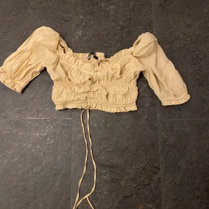 Beige croppad blus - Säljer en söt beige croppad blus med puffiga trekvartsärmar och knytband framtill. Perfekt för en somrig look eller en avslappnad dag ute. Blusen har en elastisk midja för en bekväm passform.