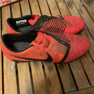 Röda Nike fotbollsskor - Säljer ett par röda Nike Phantom fotbollsskor med svarta detaljer. Skorna har en strömlinjeformad design med snörning och är perfekta för fotbollsplanen.