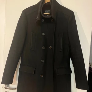 Svart rock från Holmes - Elegant svart rock från Holmes med dubbelknäppning och hög krage. Rocken har långa ärmar och två framfickor med lock. Perfekt för kyligare dagar med stil.