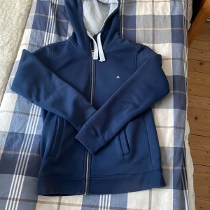 Blå huvtröja från J.Lindeberg - Snygg blå huvtröja från J.Lindeberg med dragkedja och justerbar huva. Perfekt för en avslappnad stil. Tröjan har långa ärmar och ribbade muddar för extra komfort.