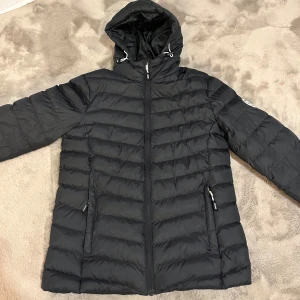 Svart dunjacka från Vinson Polo Club - En svart dunjacka från Vinson Polo Club med huva och dragkedja. Jackan har ett stilrent quiltat mönster och praktiska fickor med dragkedjor. Använd några gånger men i jätte bra skick!!