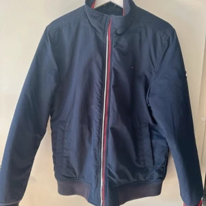 Mörkblå jacka från Tommy Hilfiger Denim - Snygg mörkblå jacka från Tommy Hilfiger Denim med dragkedja i rött, vitt och blått. Jackan har en klassisk design med ribbade muddar och en liten logotyp på ärmen. Perfekt för en stilren look.