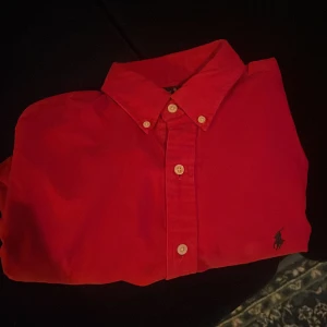Röd skjorta från Ralph Lauren - Snygg röd skjorta från Ralph Lauren med klassisk krage och knappar framtill. Den har en broderad logga på bröstet och långa ärmar. Perfekt för en stilren look.