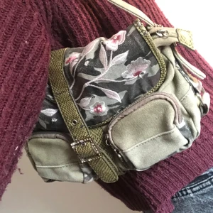 Y2K mcbling handväska - Snygg grön axelväska med broderade blommor och flera praktiska fickor. Väskan har en justerbar axelrem och stängs med ett spänne. Väskan är i ok använt skick och har skavanker. Till exempel har en del till dragkedjan lossnat (den ligger i väskan) och väskan är lite nopprig på insidan. Säljer på grund av att jag inte har fått någon användning av väskan.